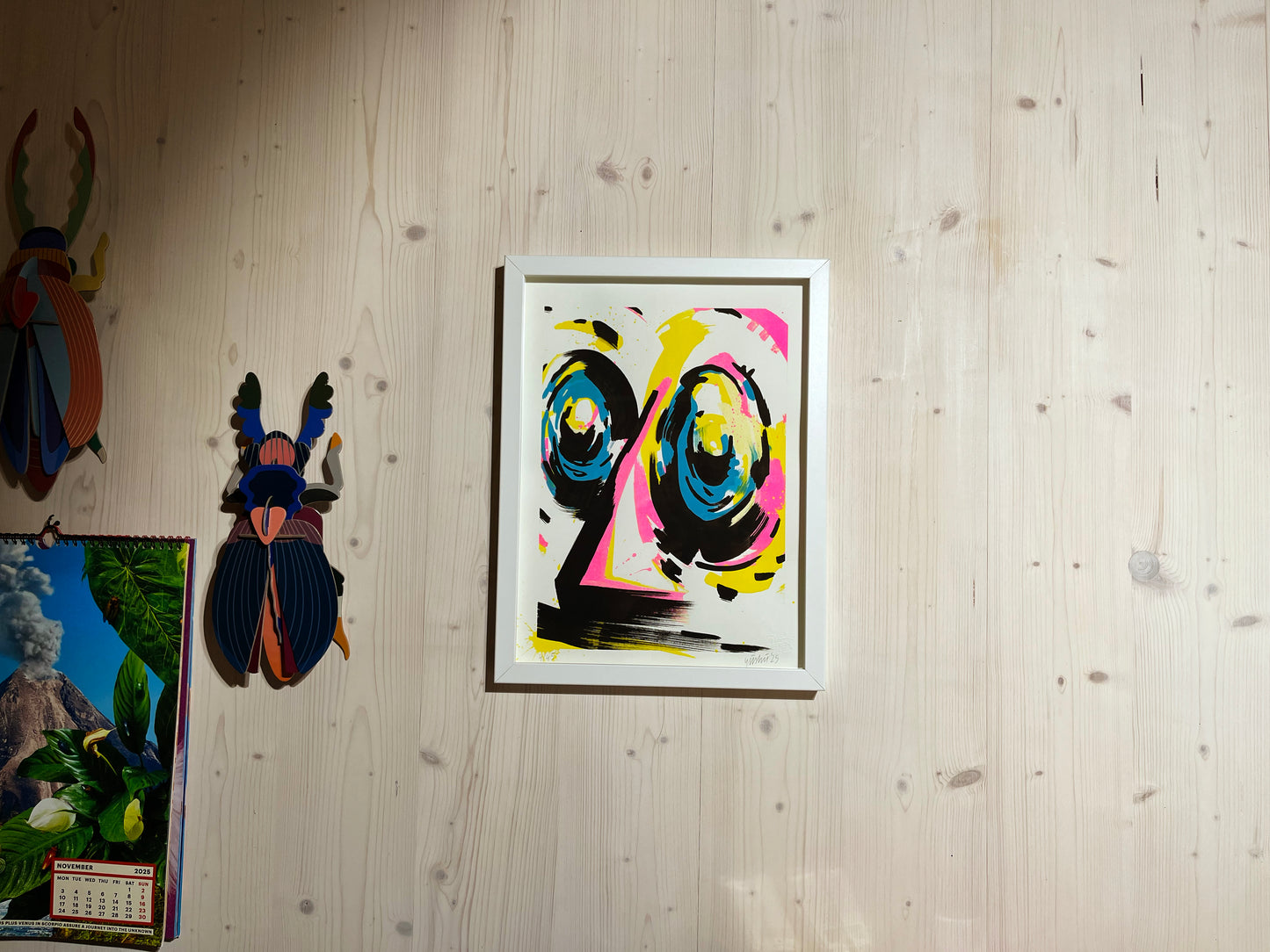 Handfinished Riso Print, Acrylmarker in 40x30 cm, gerahmt in einfachem weißen Objektrahmen. Im Atelier fotografiert. Der Rahmen hängt an einer Holzwand, in moderner Architektur. Daneben hängen Käfer von Studio Roof und der Toiletpaper Kalender. Das Bild zeigt ein abstraktes Gesicht in den Farben Cyan, Magenta, Gelb und Schwarz. Das Kunstwerk ist signiert und nummeriert von yushu – oberaffenstyle