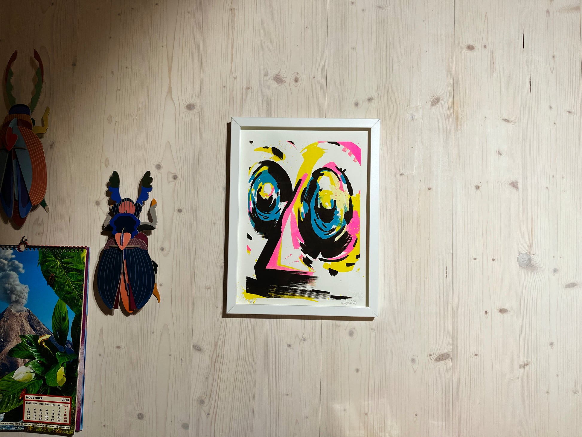 Handfinished Riso Print, Acrylmarker in 40x30 cm, gerahmt in einfachem weißen Objektrahmen. Im Atelier fotografiert. Der Rahmen hängt an einer Holzwand, in moderner Architektur. Daneben hängen Käfer von Studio Roof und der Toiletpaper Kalender. Das Bild zeigt ein abstraktes Gesicht in den Farben Cyan, Magenta, Gelb und Schwarz. Das Kunstwerk ist signiert und nummeriert von yushu – oberaffenstyle