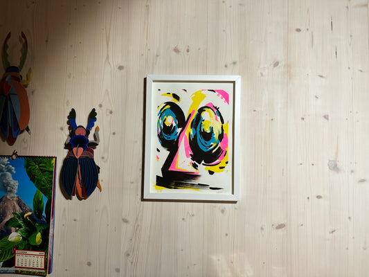 Handfinished Riso Print, Acrylmarker in 40x30 cm, gerahmt in einfachem weißen Objektrahmen. Im Atelier fotografiert. Der Rahmen hängt an einer Holzwand, in moderner Architektur. Daneben hängen Käfer von Studio Roof und der Toiletpaper Kalender. Das Bild zeigt ein abstraktes Gesicht in den Farben Cyan, Magenta, Gelb und Schwarz. Das Kunstwerk ist signiert und nummeriert von yushu – oberaffenstyle