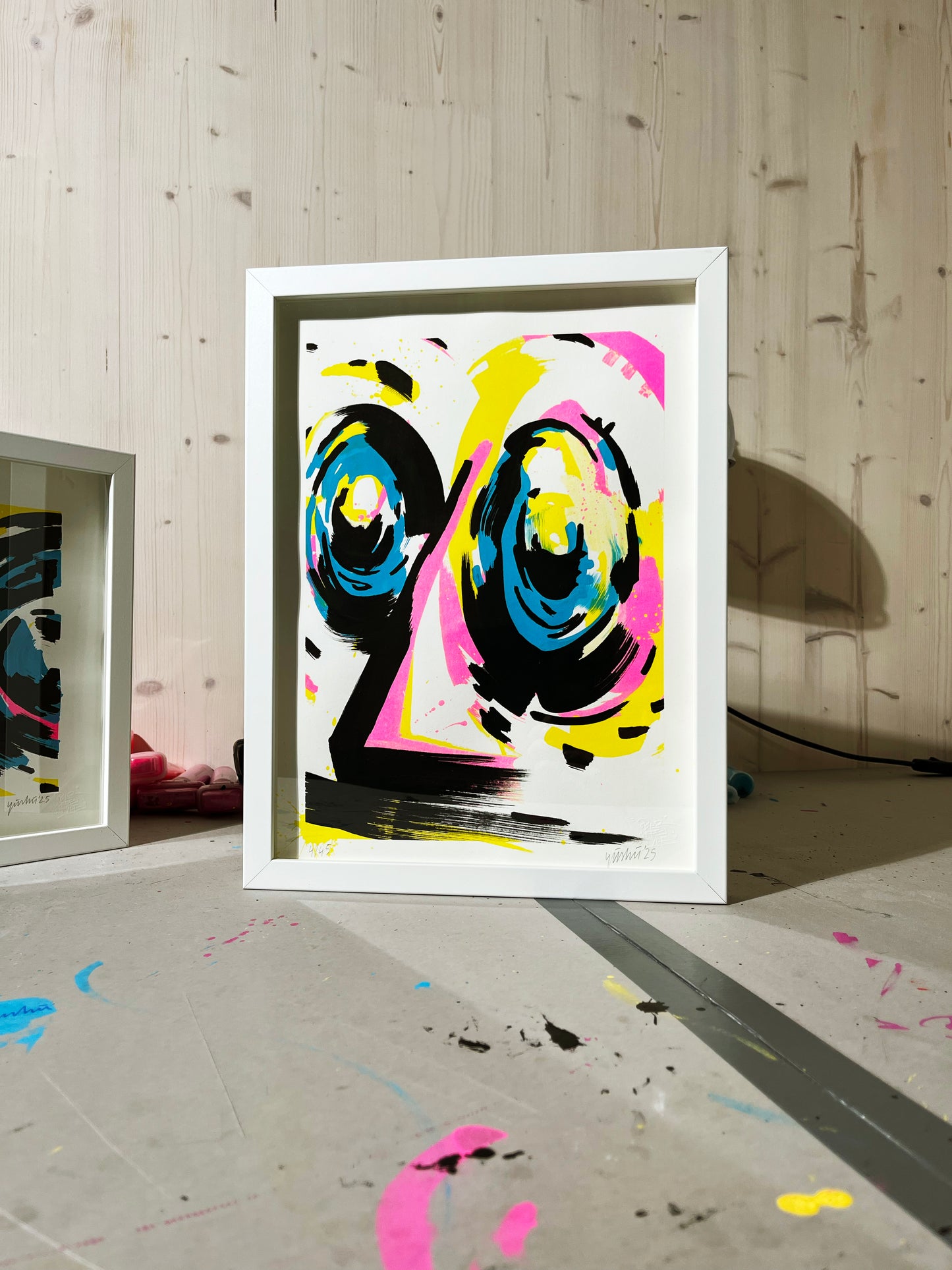 Handfinished Riso Print, 40x30 cm, gerahmt in einfachem weißen Objektrahmen im Atelier fotografiert. Der Rahmen steht auf vor einer Holzwand, in modernem Interieur. Das Bild zeigt ein abstraktes Gesicht in den Farben Cyan, Magenta, Gelb und Schwarz. Das Kunstwerk ist signiert und nummeriert. 